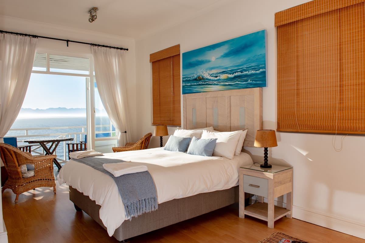 Un dormitorio con una cama grande, una mesita de noche, una silla de mimbre y vistas al océano, además de un cuadro marino.