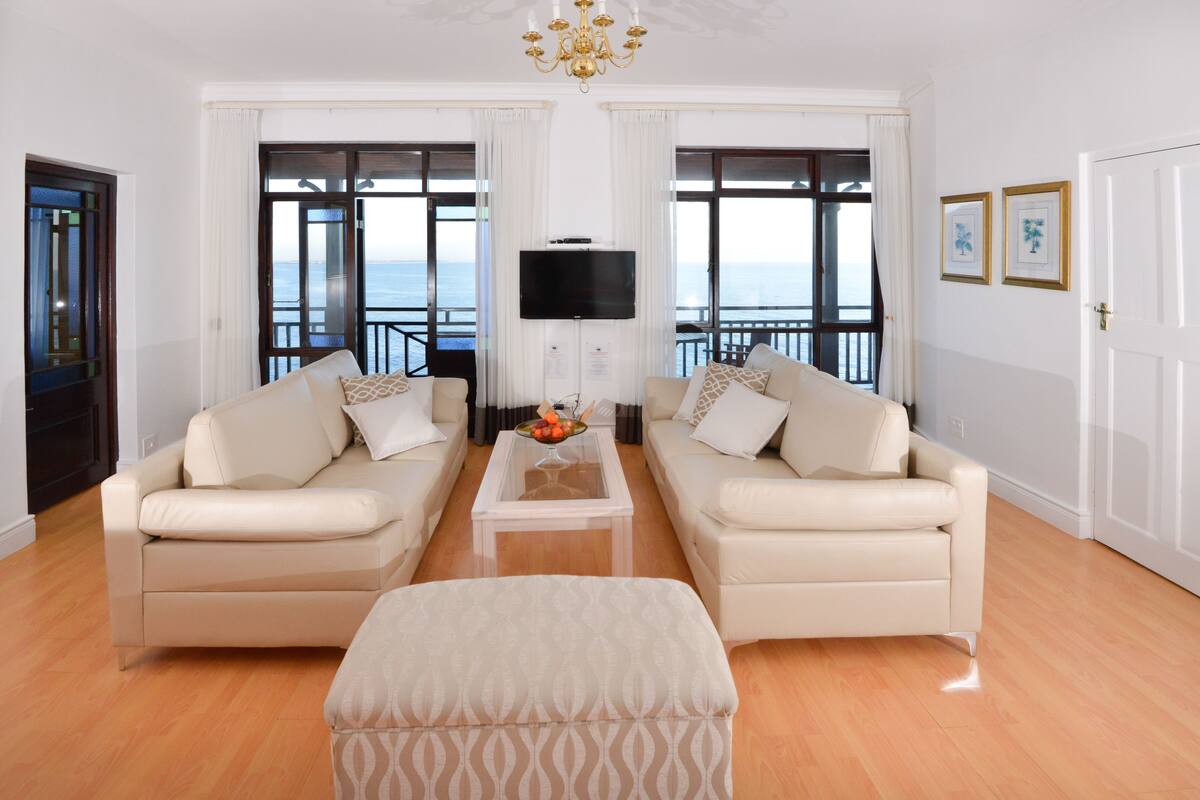 Una sala de estar con dos sofás, una mesa de centro y un televisor. Hay ventanas amplias con vista al mar.