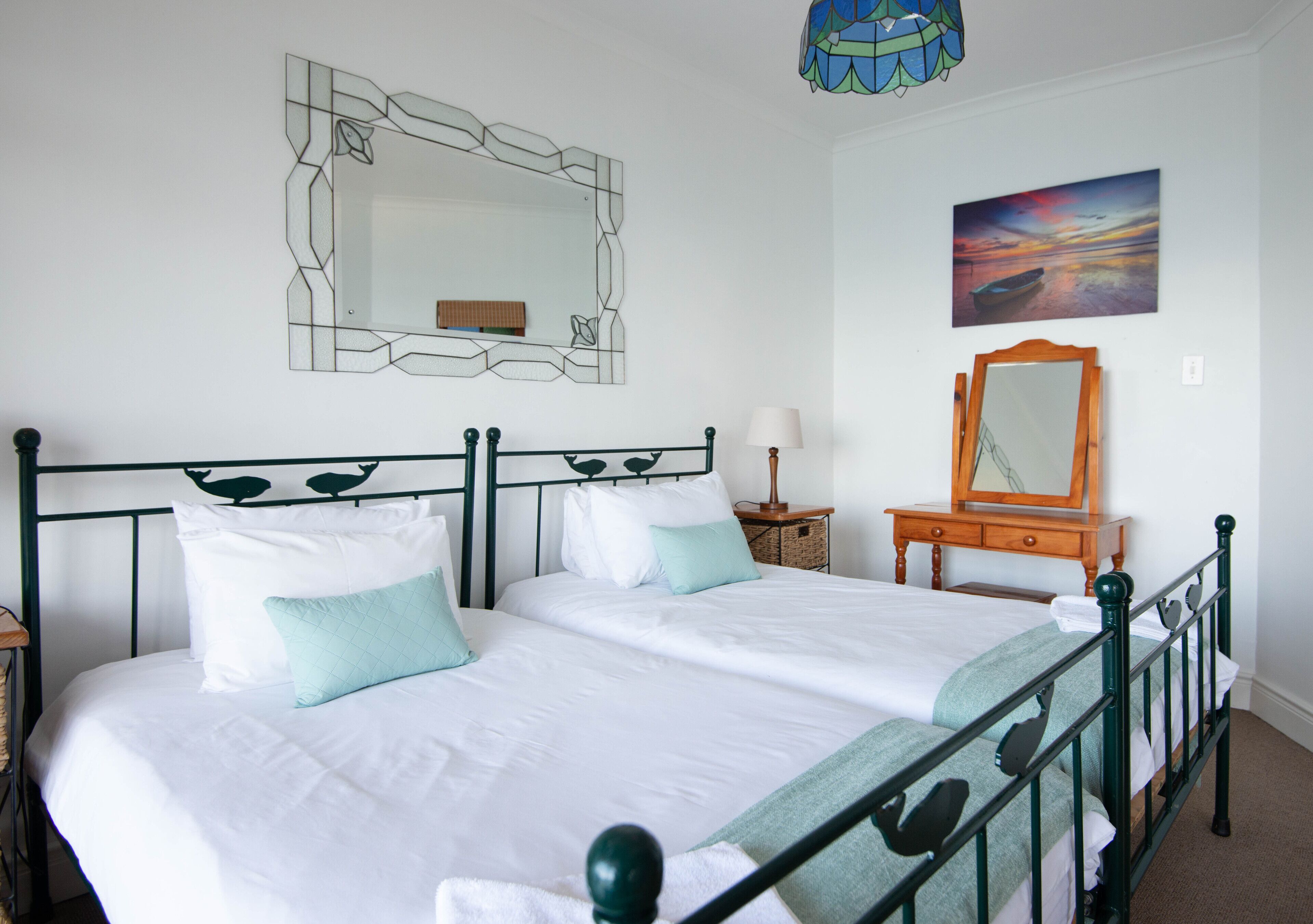 A double bed with white bedding and teal pillows, a wooden nightstand with a lamp, a mirror, and a framed abstract painting on the wall.