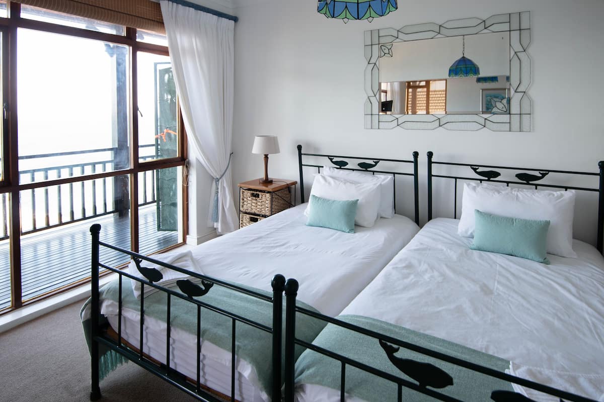 Una cama doble con estructura metálica, dos almohadas, una mesita de noche con lámpara y un ventanal con cortinas.