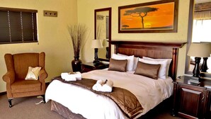 A hotel room with a large bed, a wooden nightstand, a chair, a lamp, and a painting of a tree. - Almar Exclusive Game Ranch (Bloemhof)