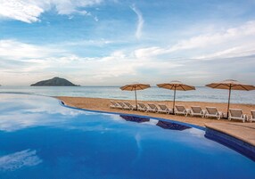 Zona junto a la piscina con sillones de descanso y sombrillas, con vista a la playa y el mar.
