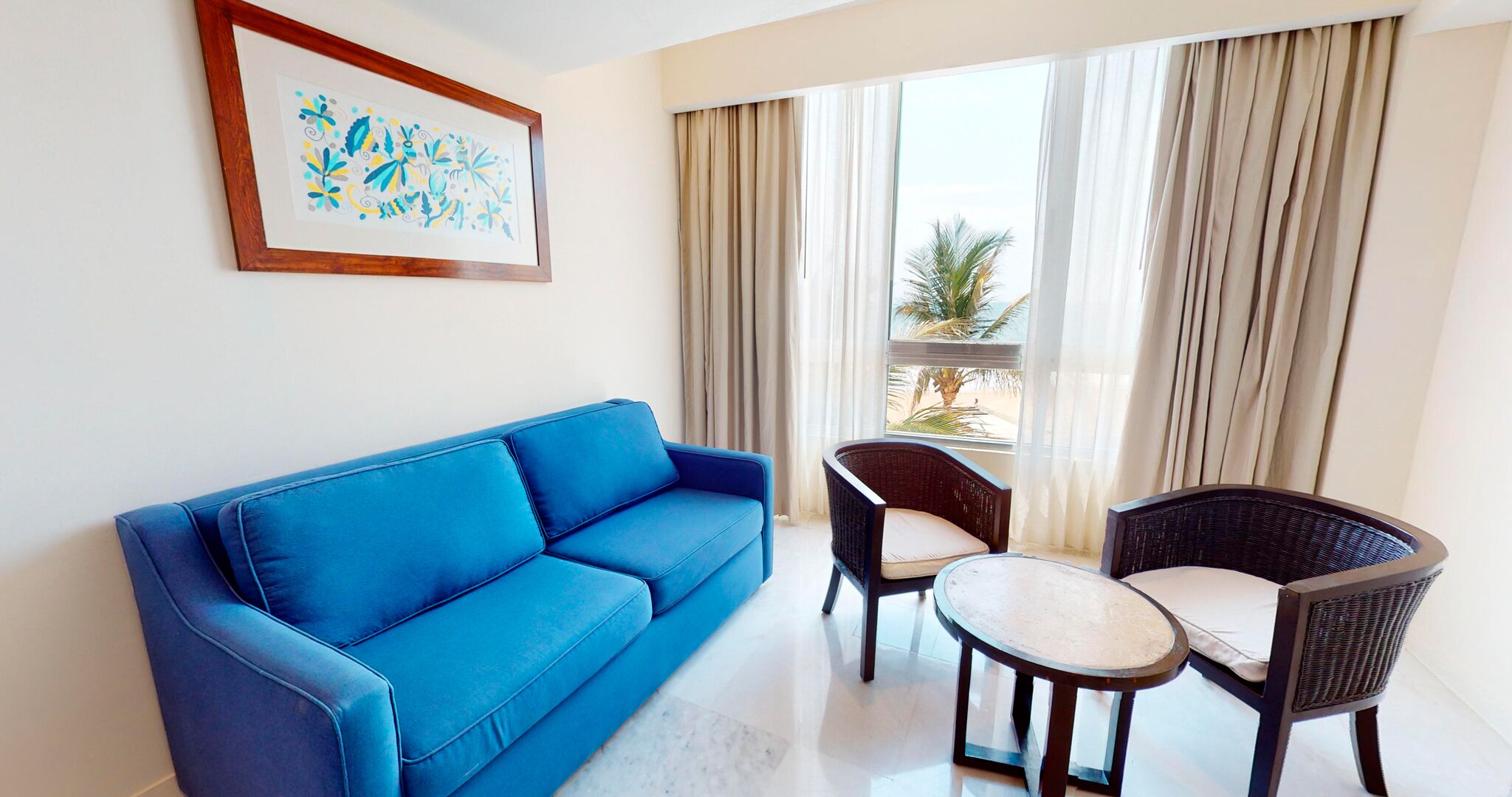 A room with a blue sofa, two wicker chairs, a small round table, a framed artwork, and a view of palm trees through sheer curtains.
