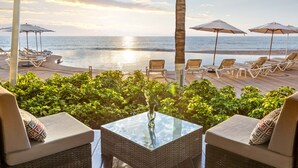Un área de descanso en la playa con mobiliario de mimbre, una mesa de vidrio y sombrillas.