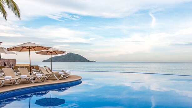 Una piscina infinita con sillas de descanso y sombrillas, con vistas al mar tranquilo y una isla distante.