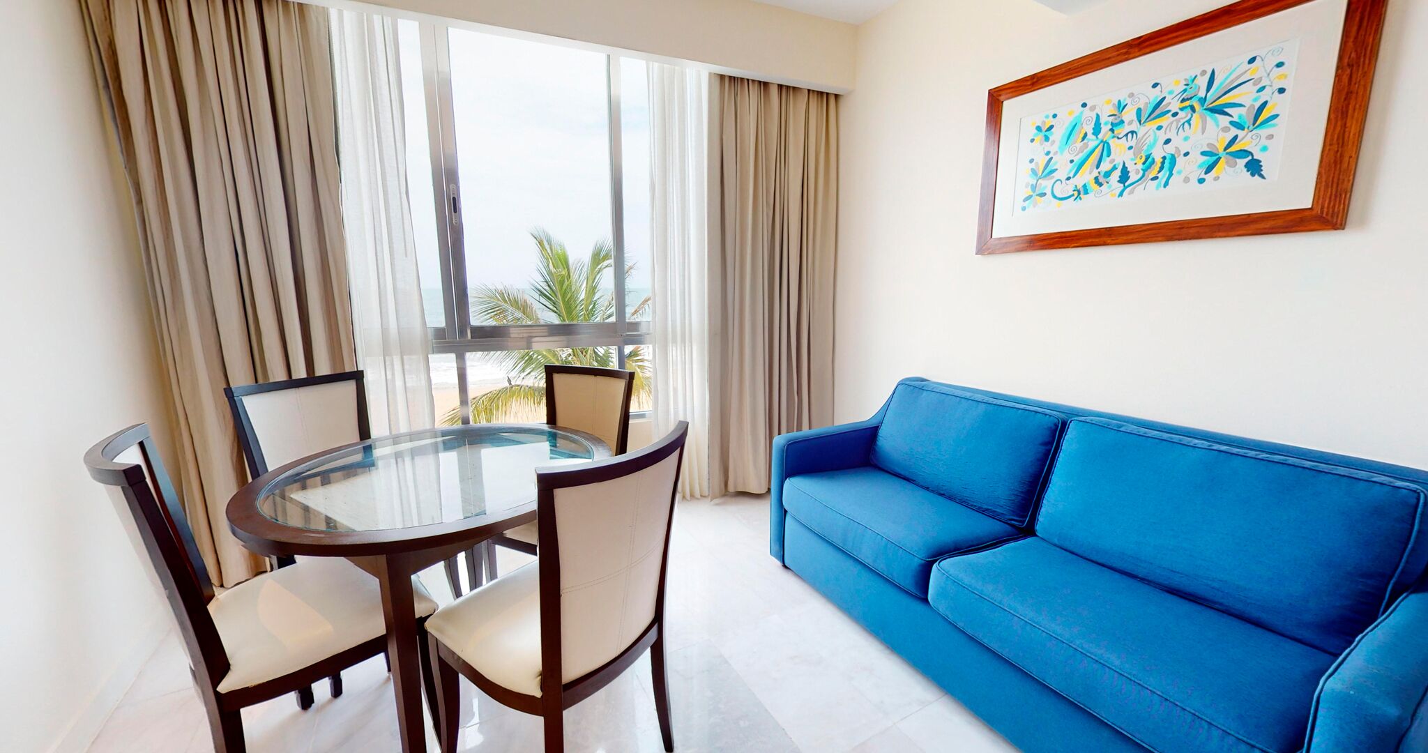 Una habitación con una mesa de vidrio, cuatro sillas, un sofá azul y un ventanal con cortinas.