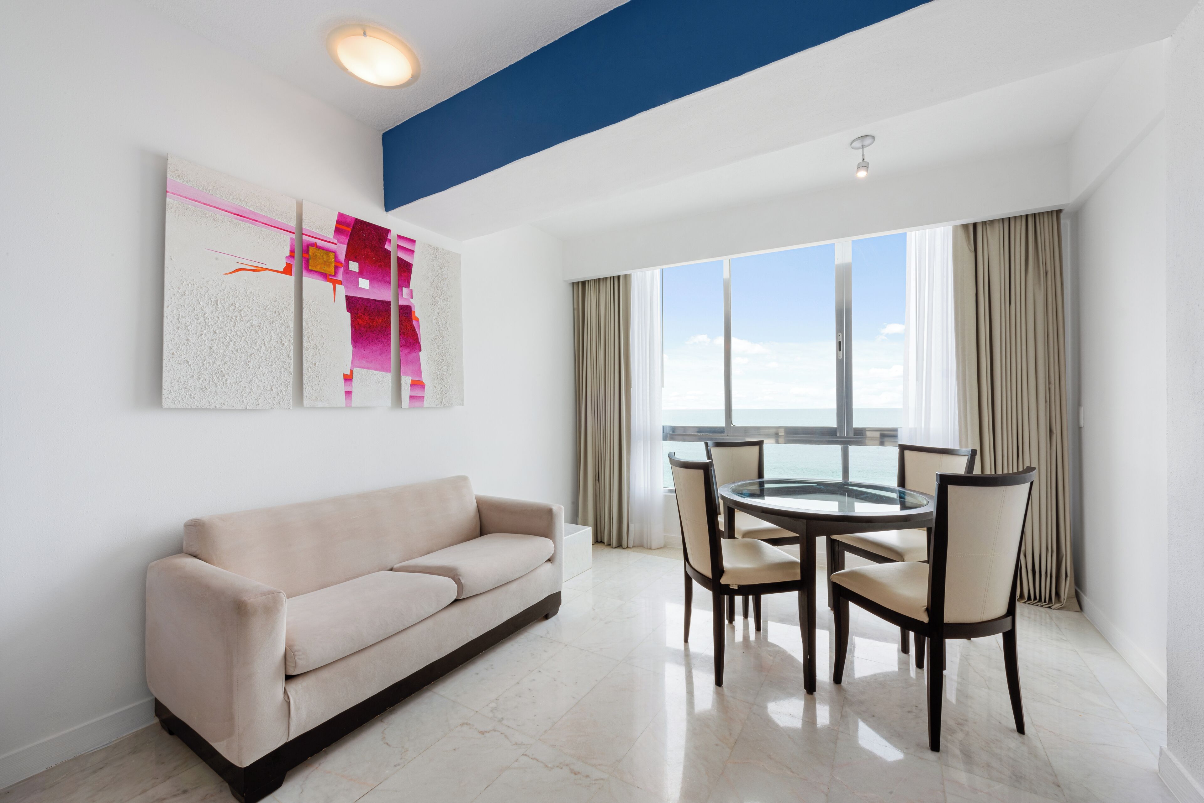 A modern hotel room with a sofa, dining table, chairs, and a large window with curtains.