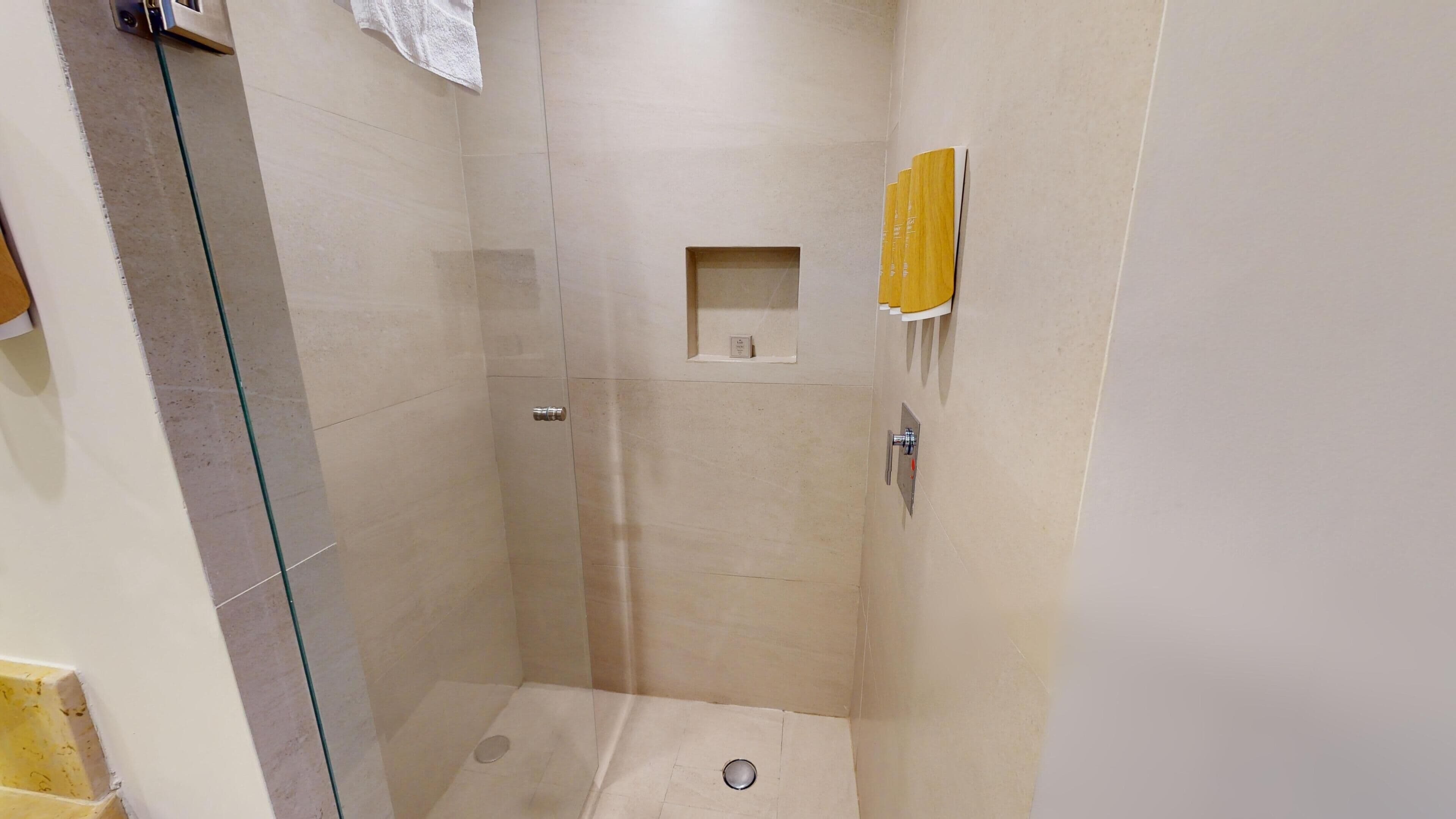 Un box de ducha pequeño con azulejos y puerta de vidrio, una toalla colgada en la pared y un estante con artículos de aseo.