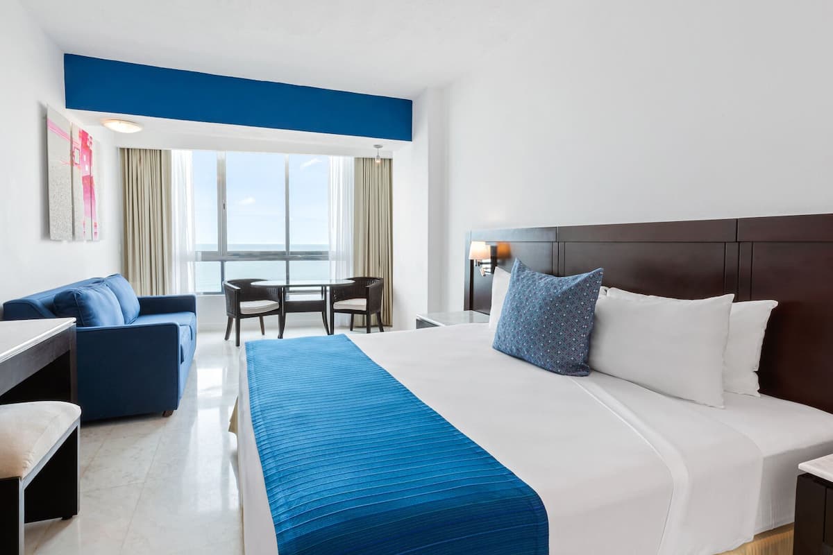 Een hotelkamer met een bed, een bureau, een stoel, een bank, een raam met gordijnen en uitzicht op zee.