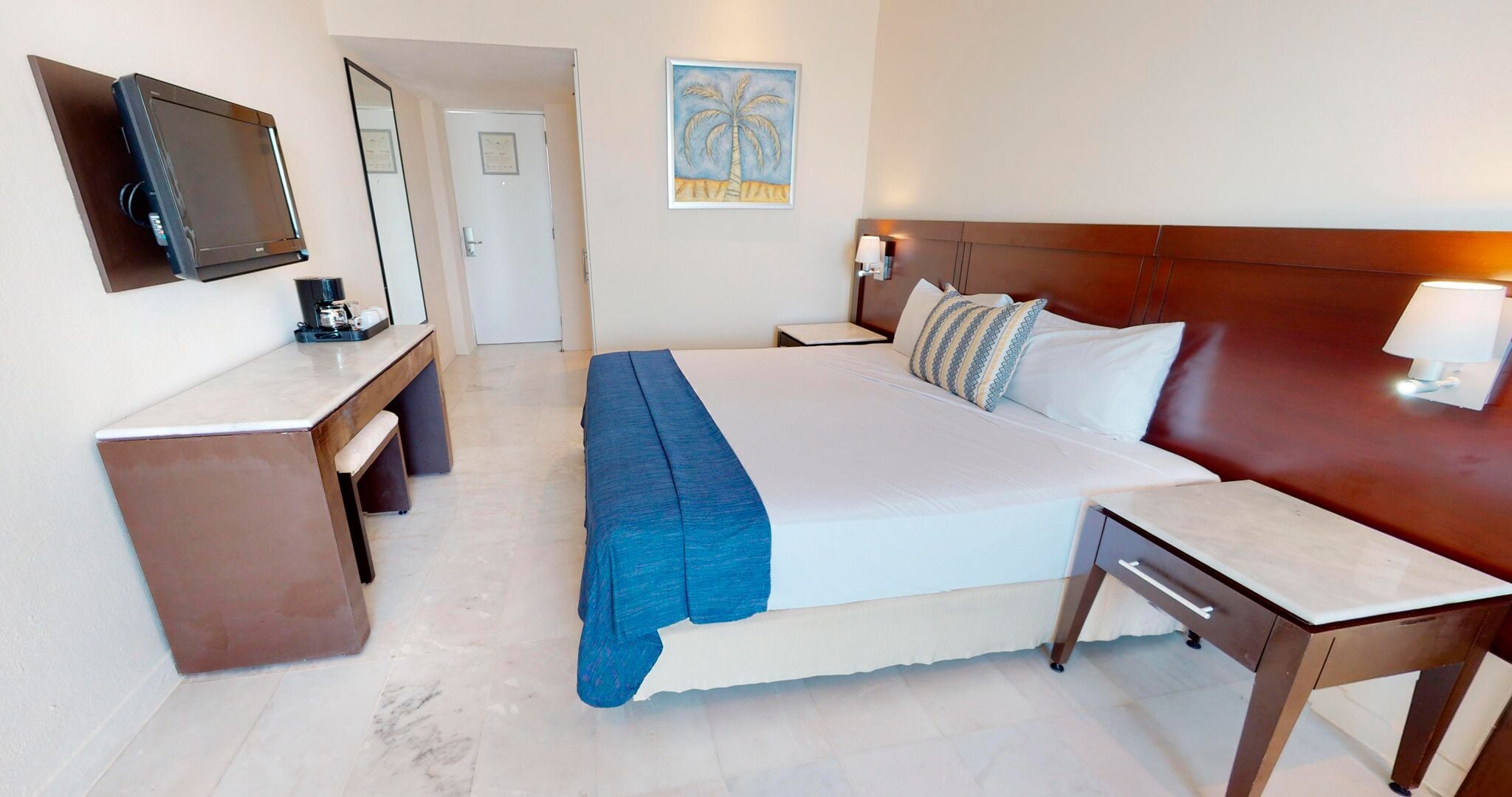 Habitación de hotel con cama, mesitas de noche, un escritorio con silla, televisor montado en la pared y una puerta.