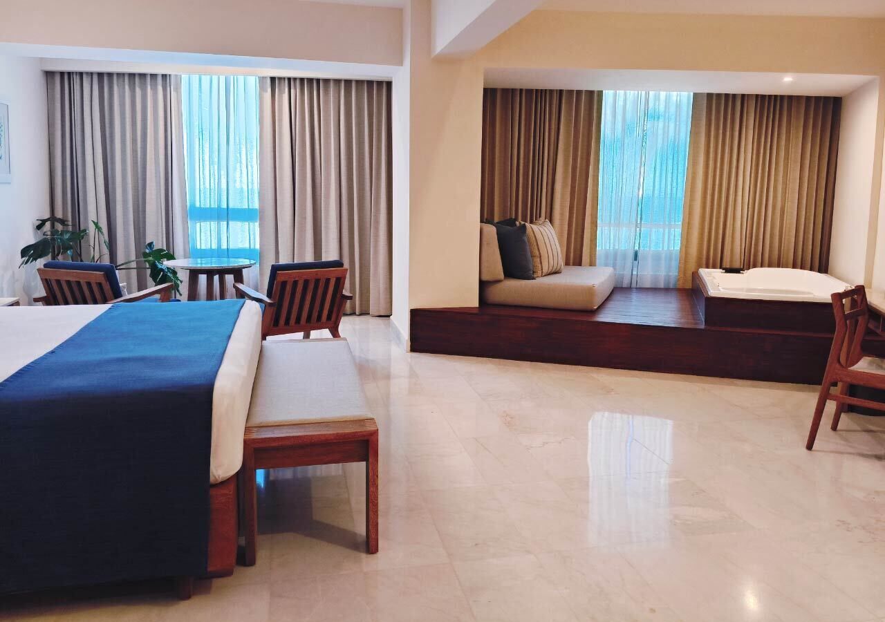 A hotel room with a large bed, a sofa, a dining table with chairs, and a view of the outdoors through large windows.