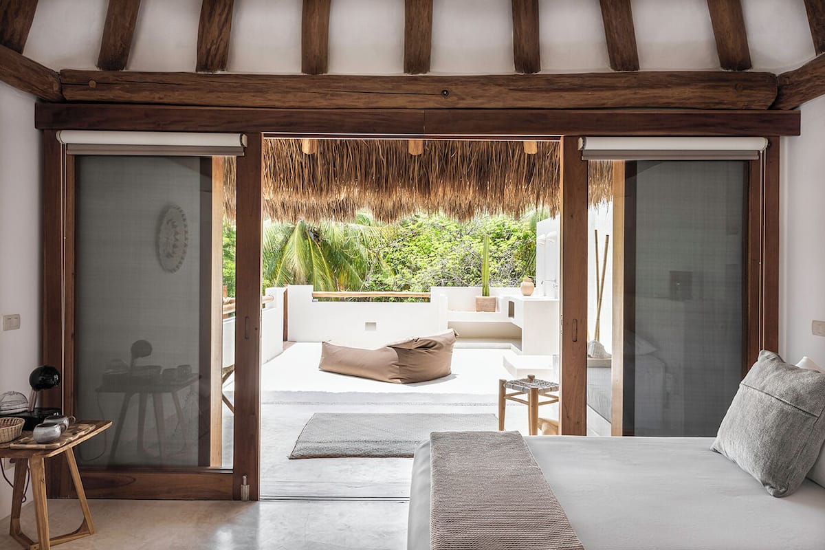 A bedroom with a large bed, a wooden table, and a view of an outdoor patio with a thatched roof and greenery.