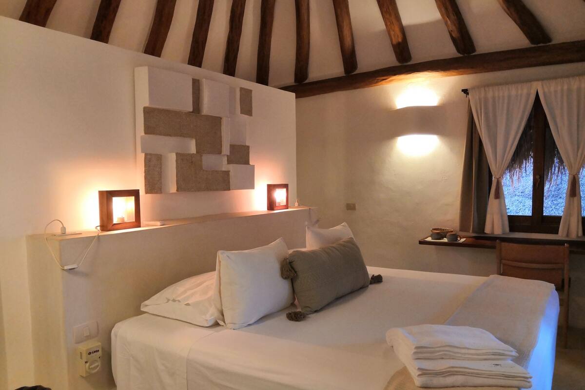 A bedroom with a bed, pillows, a bedside table, a lamp, and a window with curtains.