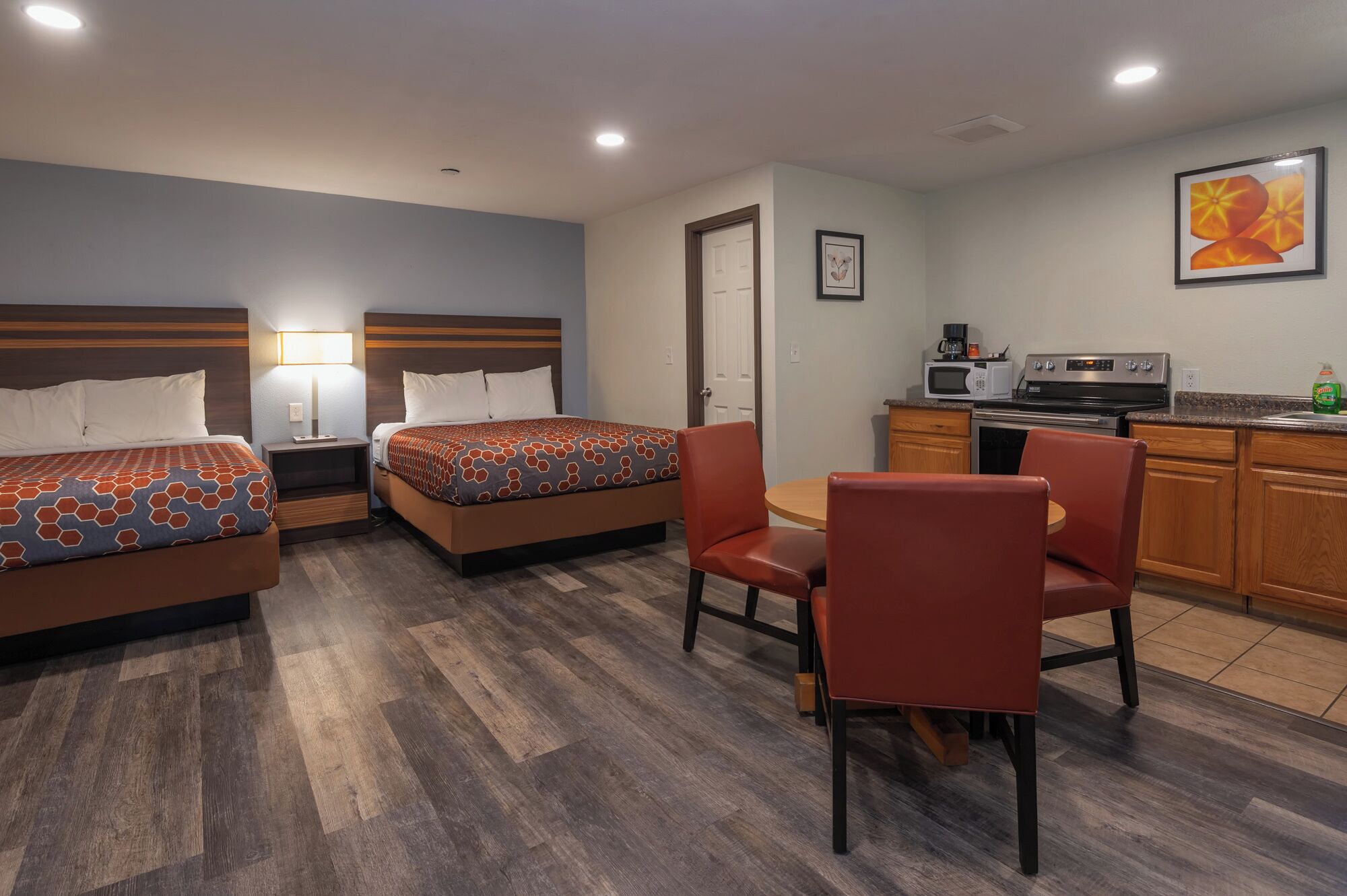 A hotel room with two beds, a dining area with chairs, a kitchen with appliances, and a door leading to another room.