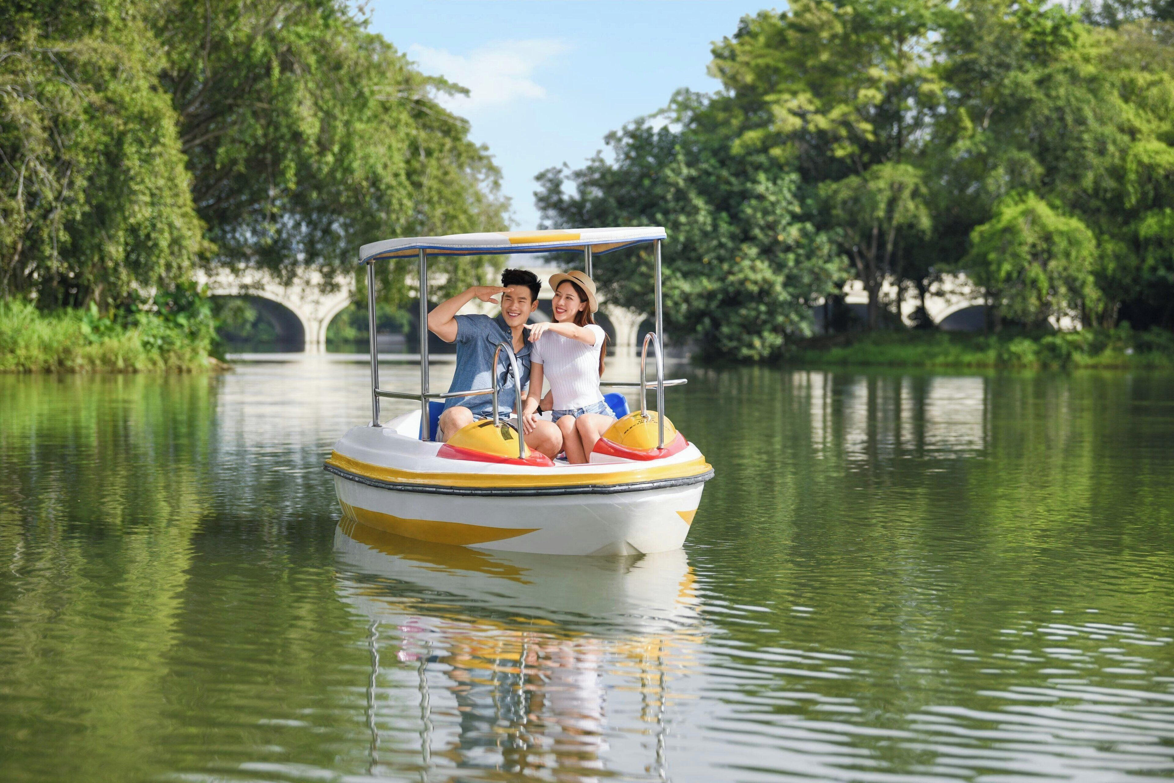 A boat with a canopy and two people on it, floating on calm water with trees and a bridge in the background.
