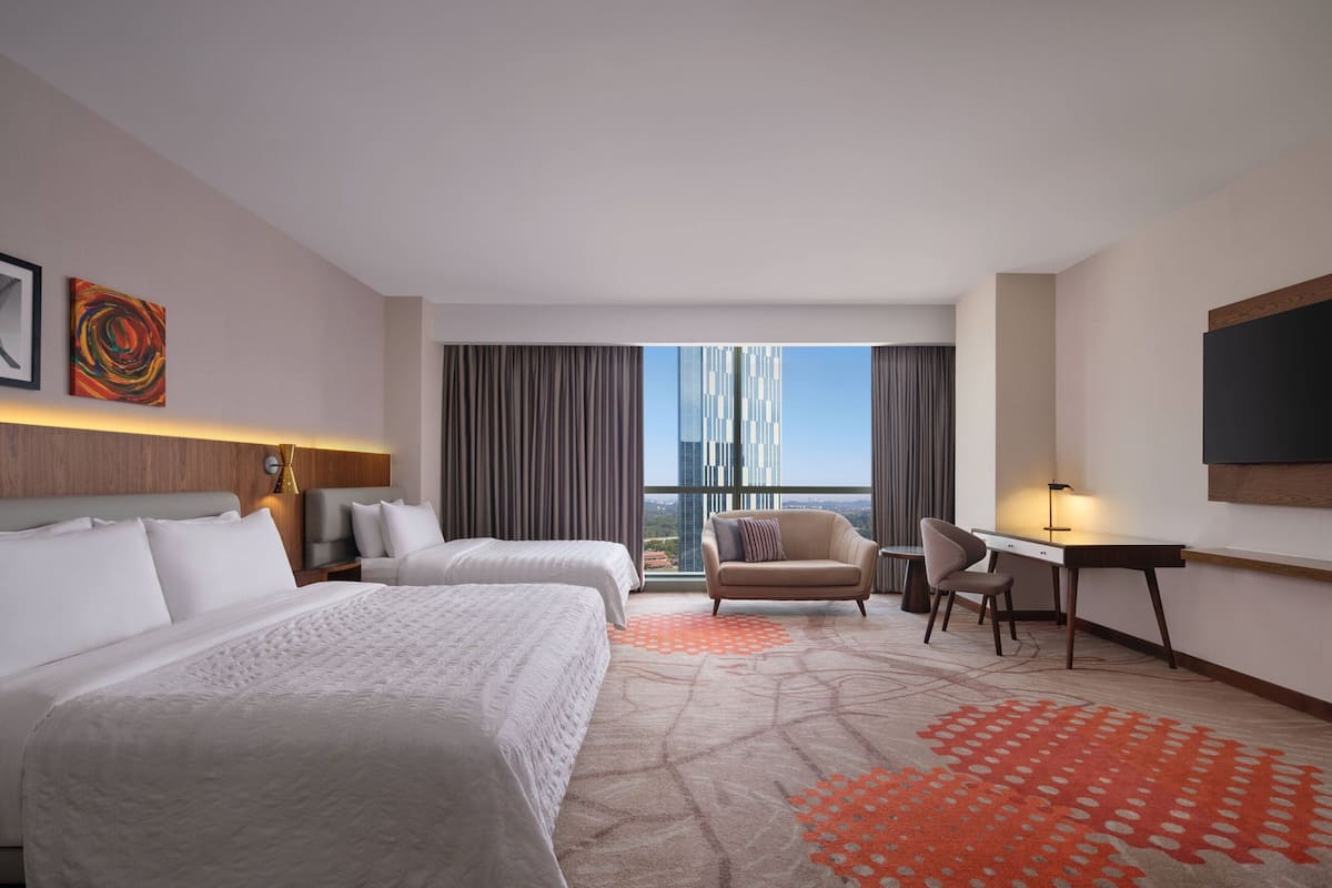 A hotel room with two beds, a desk, a chair, a TV, and a large window with a city view.