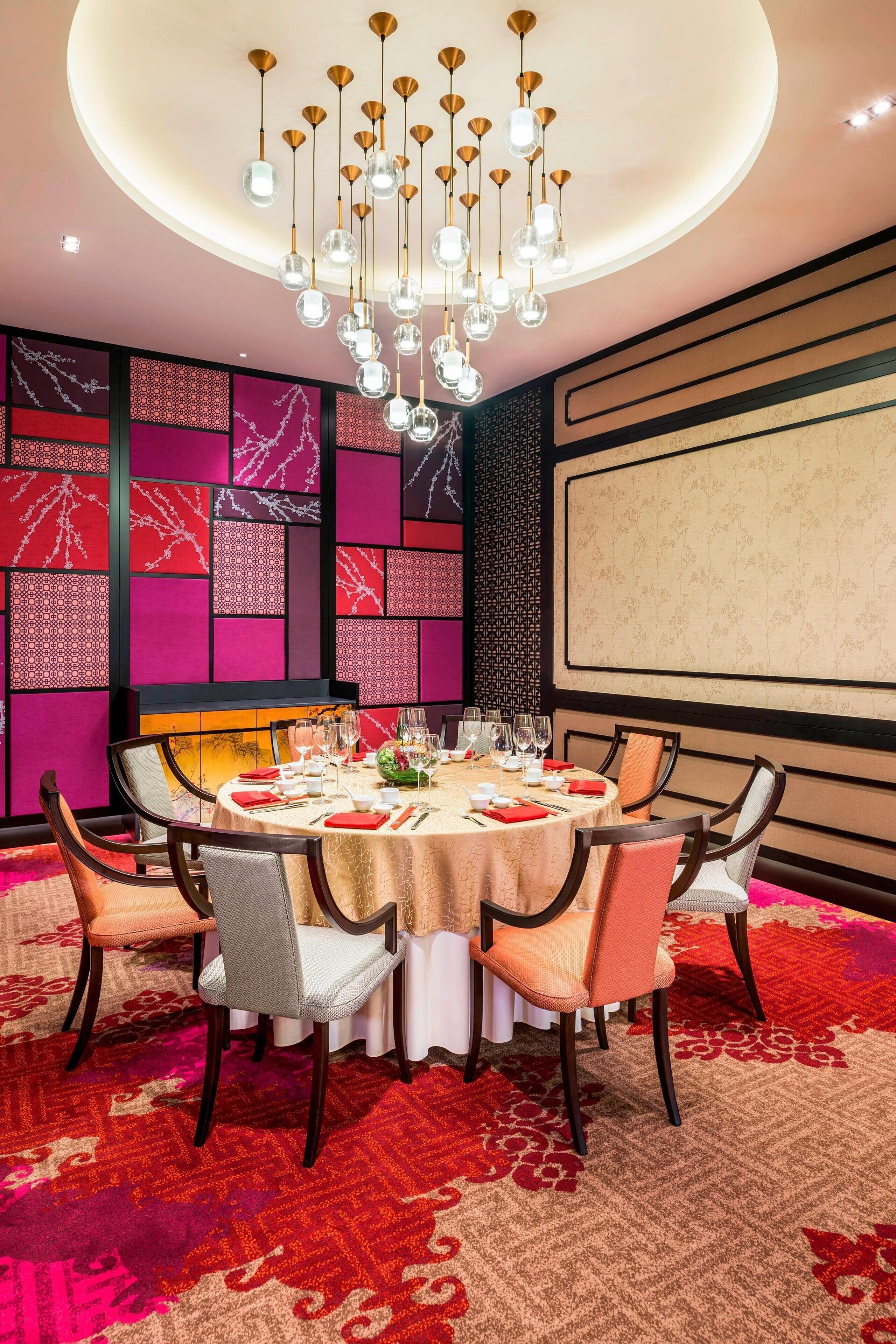 Un coin repas avec une table ronde dressée pour six personnes, entourée de chaises, d’un lustre et d’un tapis à motifs.