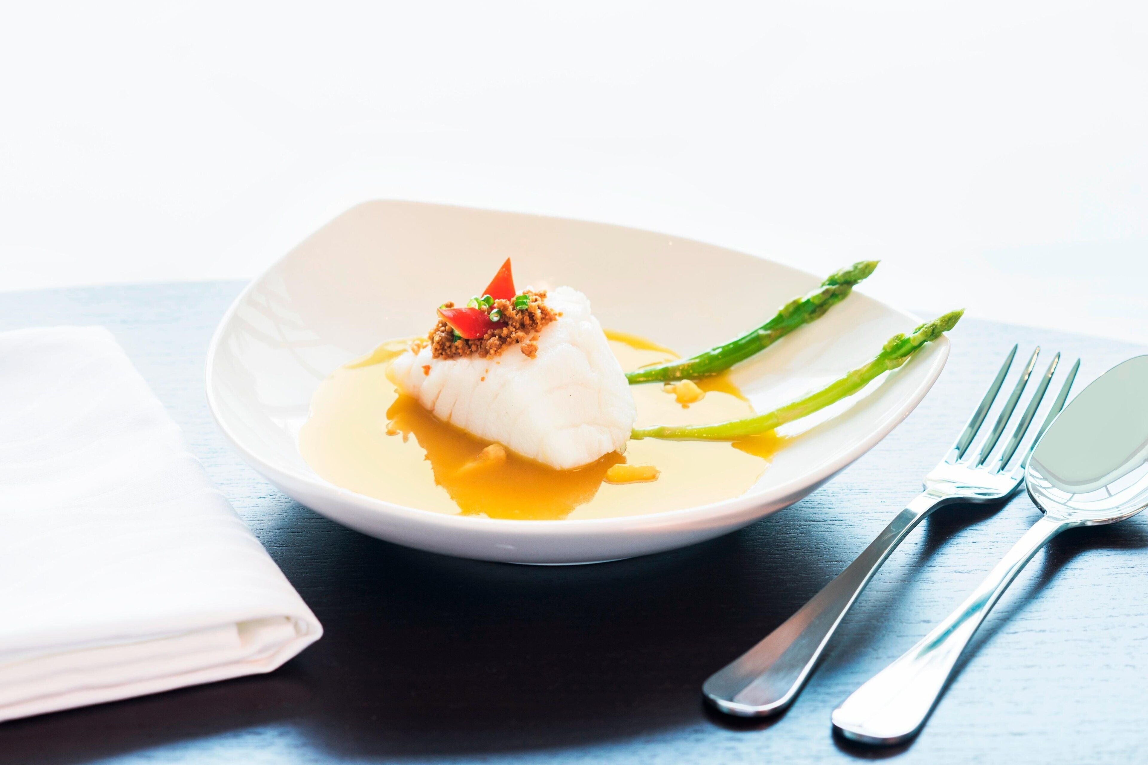 A white plate with a piece of white fish, green asparagus, and a garnish on a dark table.