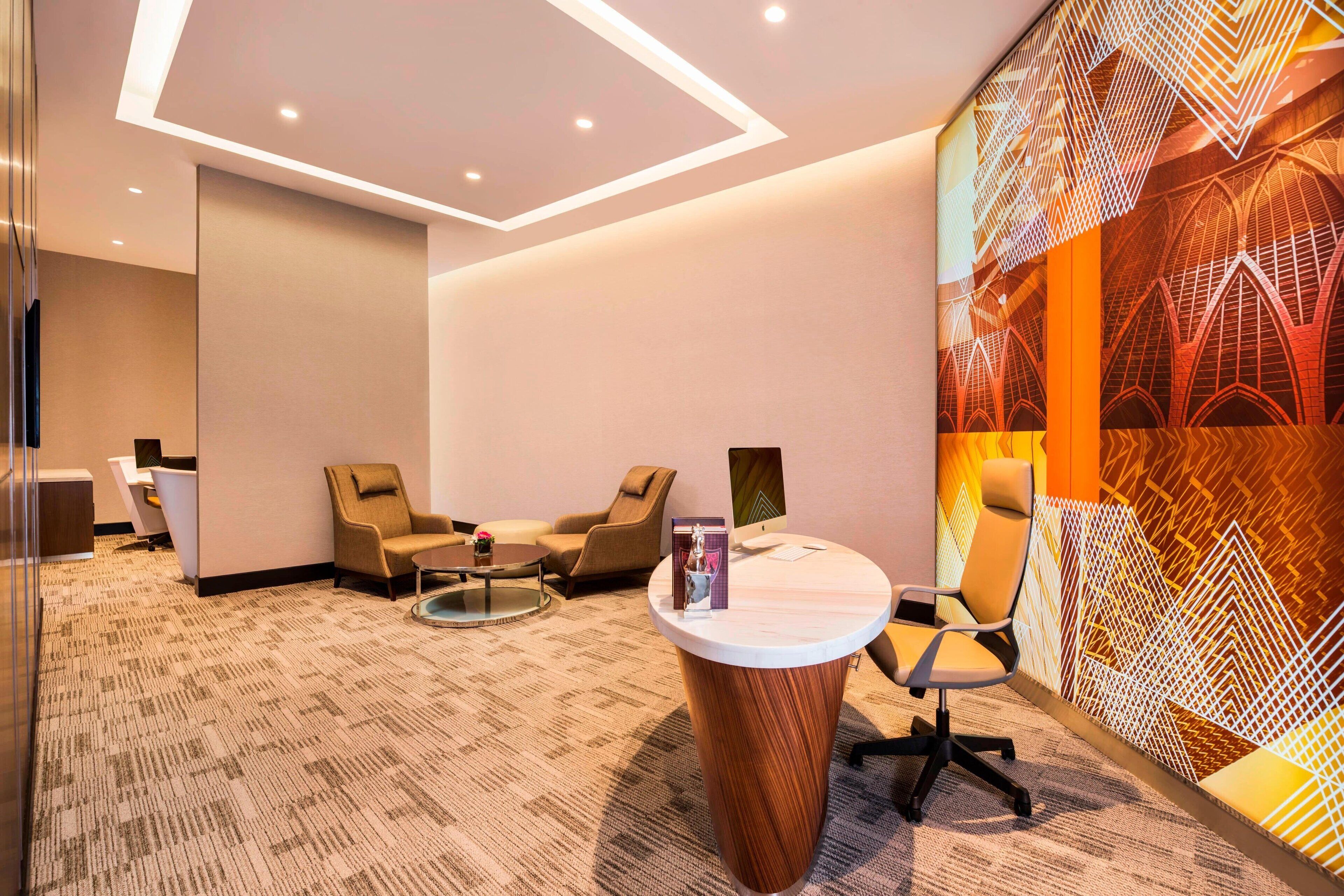 Una habitación de hotel moderna con piso alfombrado, un escritorio redondo de madera, dos sillones, un separador de vidrio y un papel tapiz estampado.