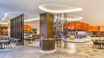 Una moderna hall d'albergo con una reception centrale, diverse aree ristorazione e una cucina sullo sfondo.