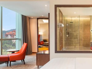 Een hotelkamer met een groot raam, een rode stoel, een klein tafeltje en een douchecabine met glazen wanden.