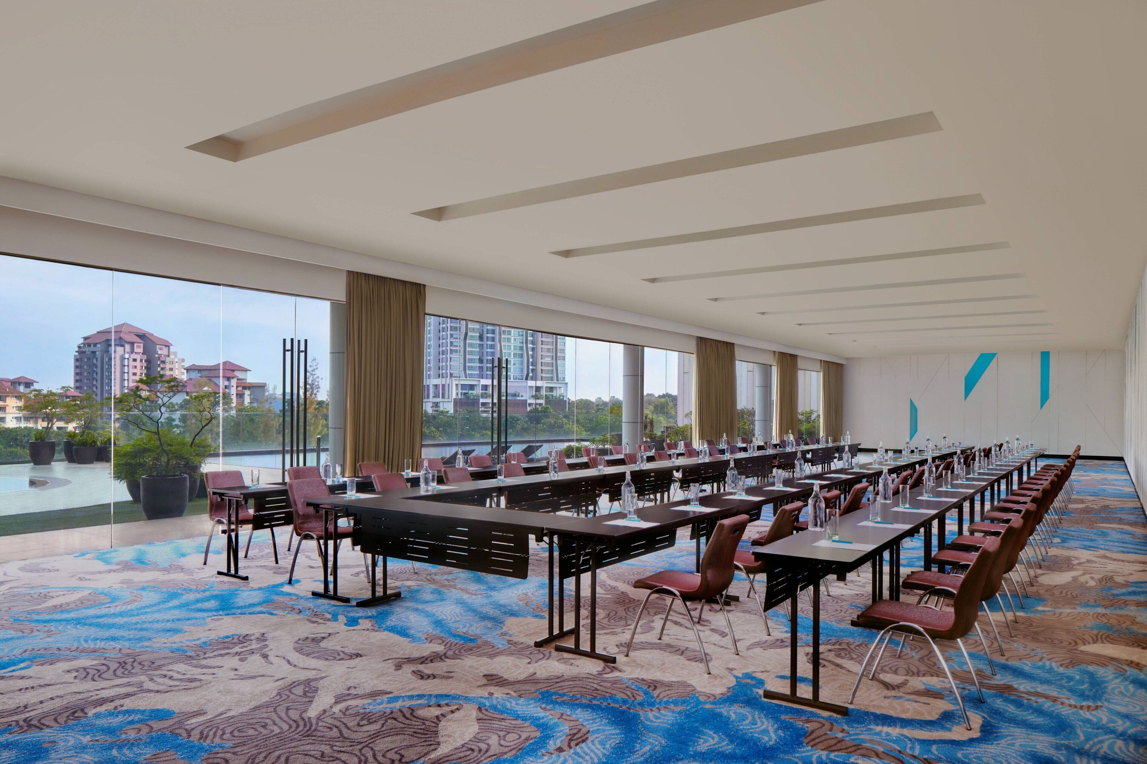 Un salón de conferencias con largas mesas y sillas, ventanas amplias y vistas a edificios.
