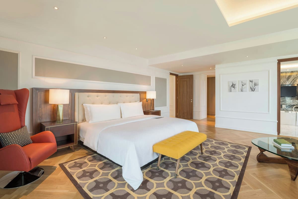 A hotel room with a large bed, a red armchair, a yellow ottoman, a glass coffee table, and a patterned rug.