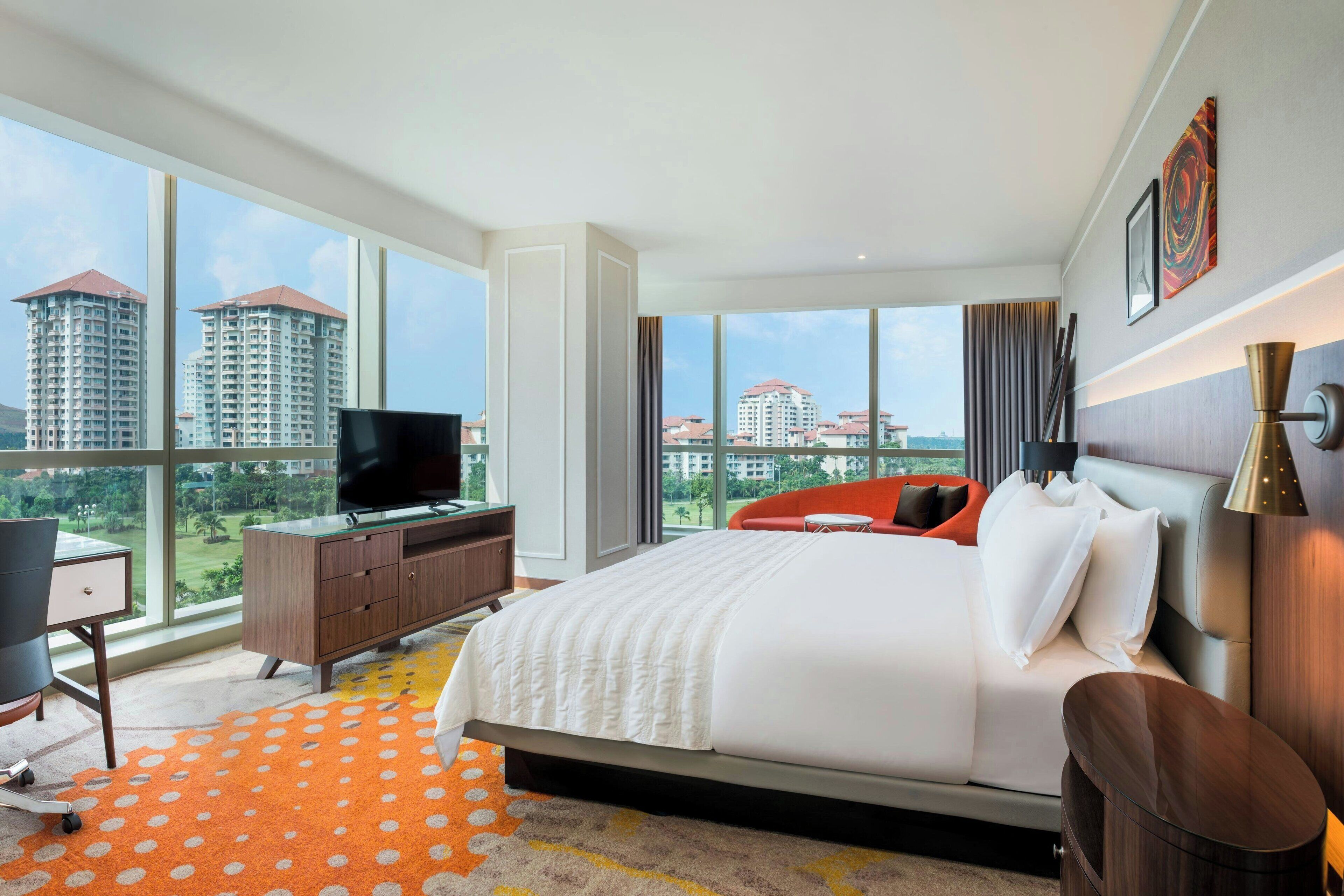 A modern hotel room with a large bed, a desk with a chair, a TV, and a view of high-rise buildings.