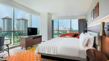 A modern hotel room with a large bed, a desk with a chair, a TV, and a view of high-rise buildings.