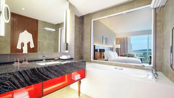 Un baño moderno con un espejo grande, encimera de mármol, bañera blanca y vistas a un dormitorio con cama y paisaje urbano.