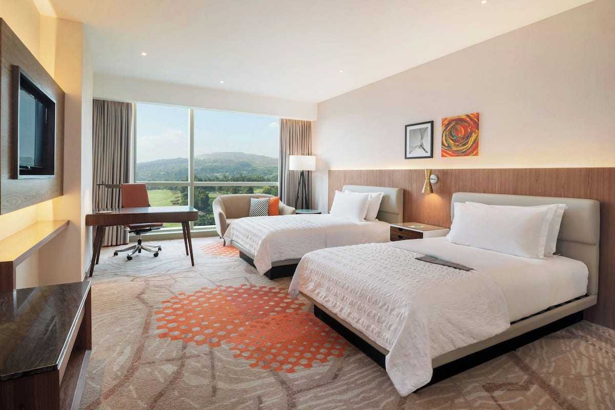 A hotel room with two beds, a desk, a TV, and a large window with a view of the countryside.