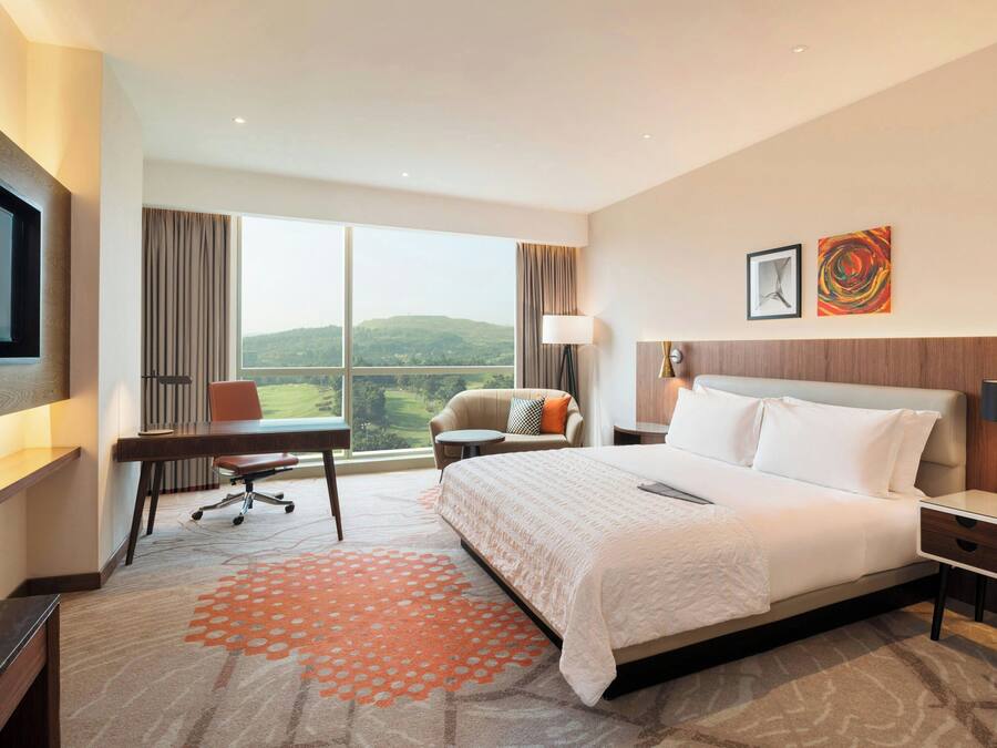 A hotel room with a large bed, a desk, a chair, a TV, and a window with a view of the hills.