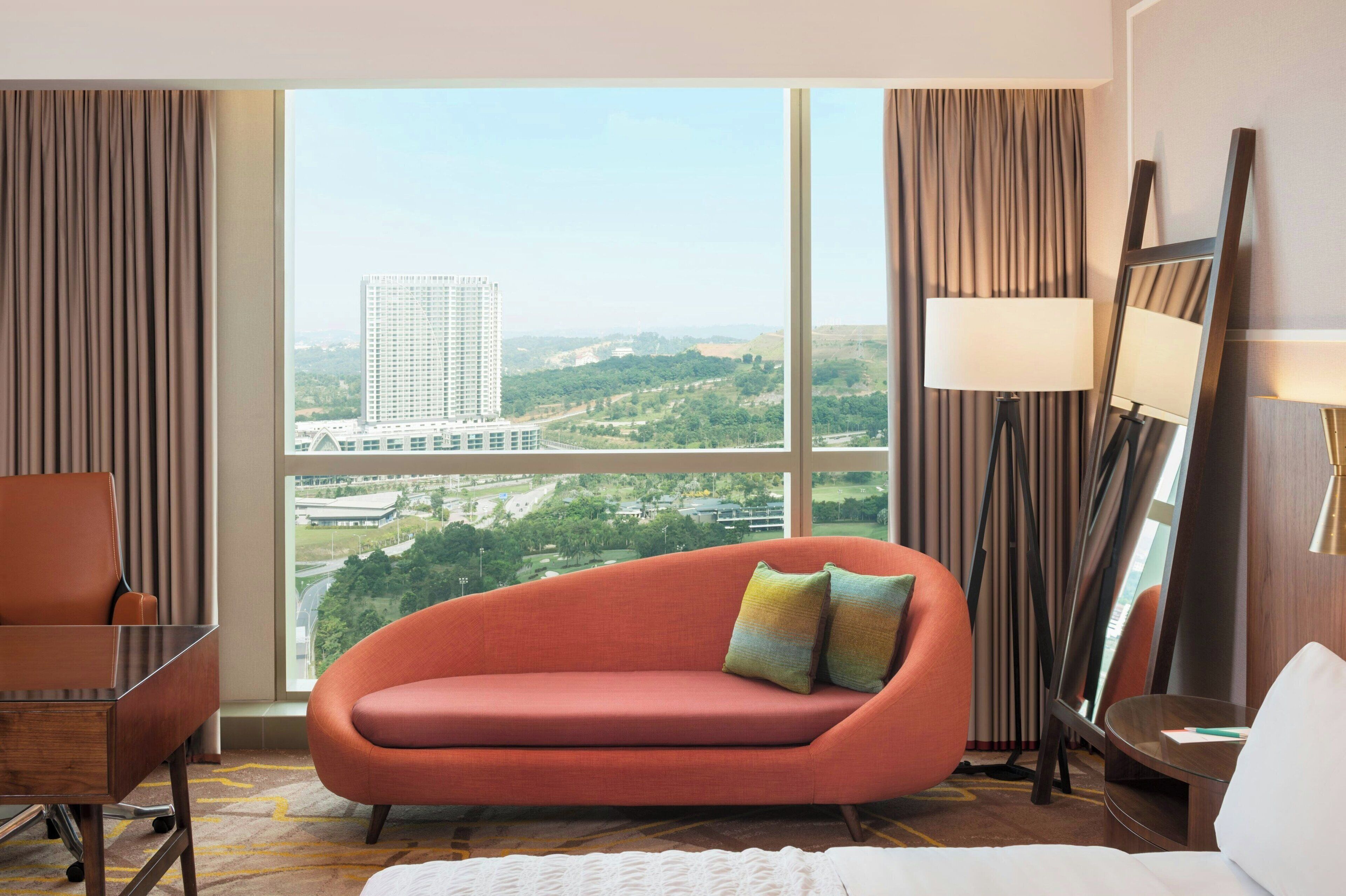 A hotel room with a large window, a sofa, a chair, a bed, and a lamp.