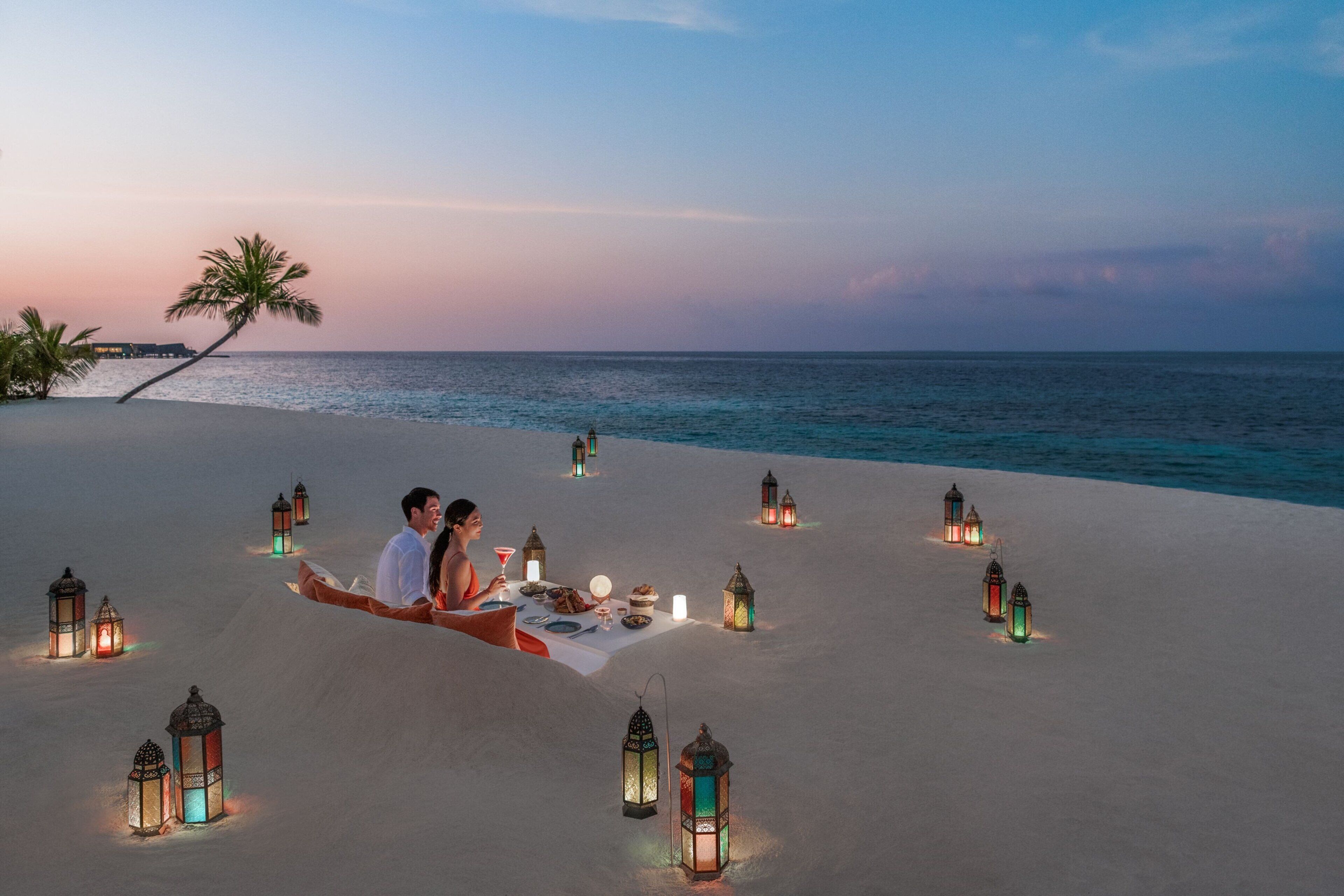 A couple dining on a beach with lanterns and palm trees in the background.