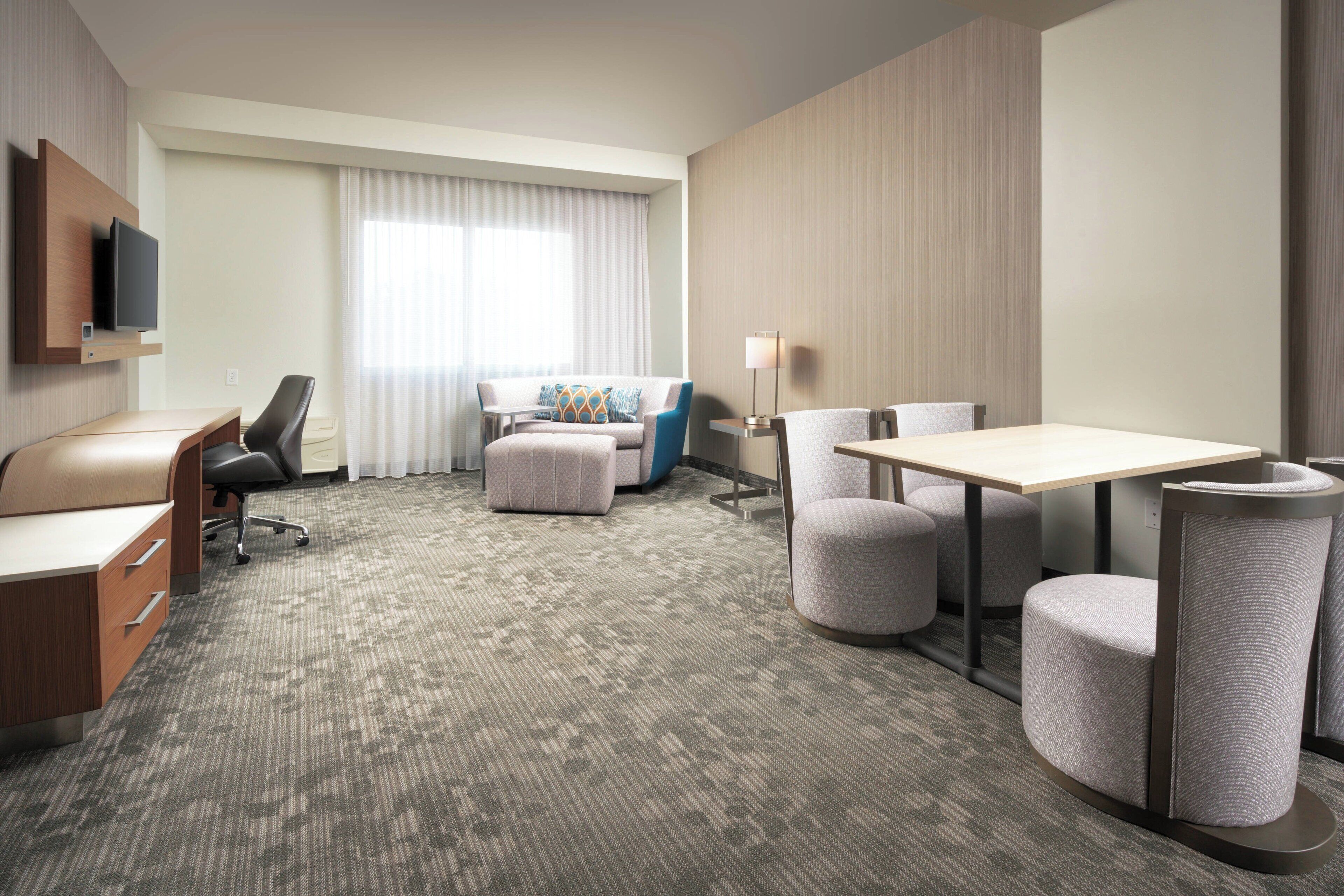 Une chambre d’hôtel équipée d’un bureau, d’une chaise, d’un pouf et d’un canapé agrémenté d’un coussin à motifs.
