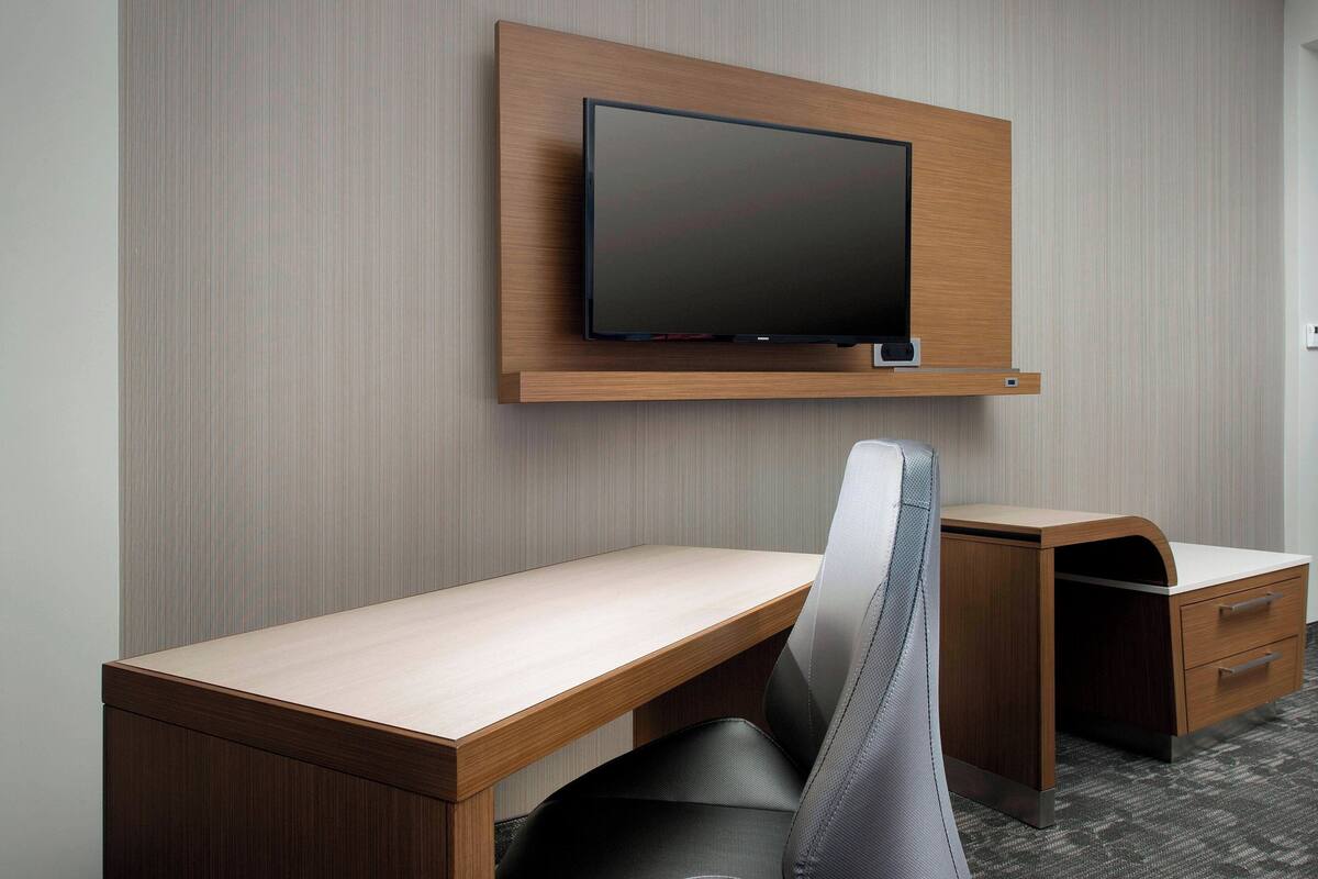 A modern hotel room with a wooden desk, a comfortable chair, a flat-screen TV mounted on the wall, and a small drawer unit.