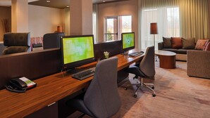 A hotel lobby with a reception desk, two computer workstations, a seating area with a sofa and armchairs, and a large window with curtains.