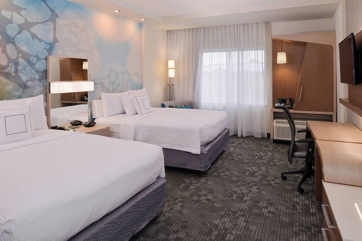 A hotel room with two beds, a desk, a TV, and a large window with curtains.