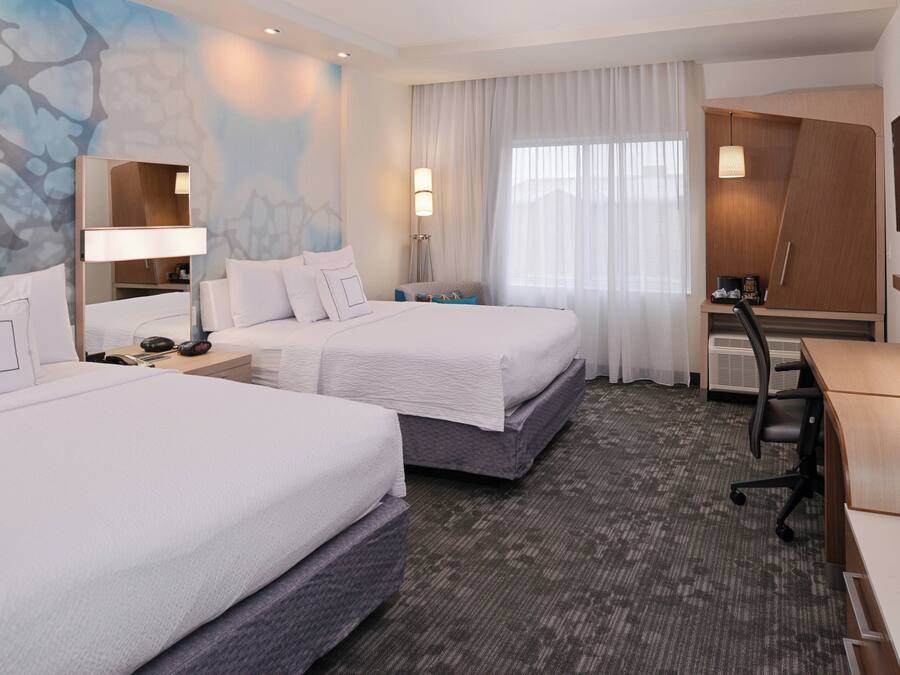 Habitación de hotel con dos camas, un escritorio, un televisor y un ventanal con cortinas.