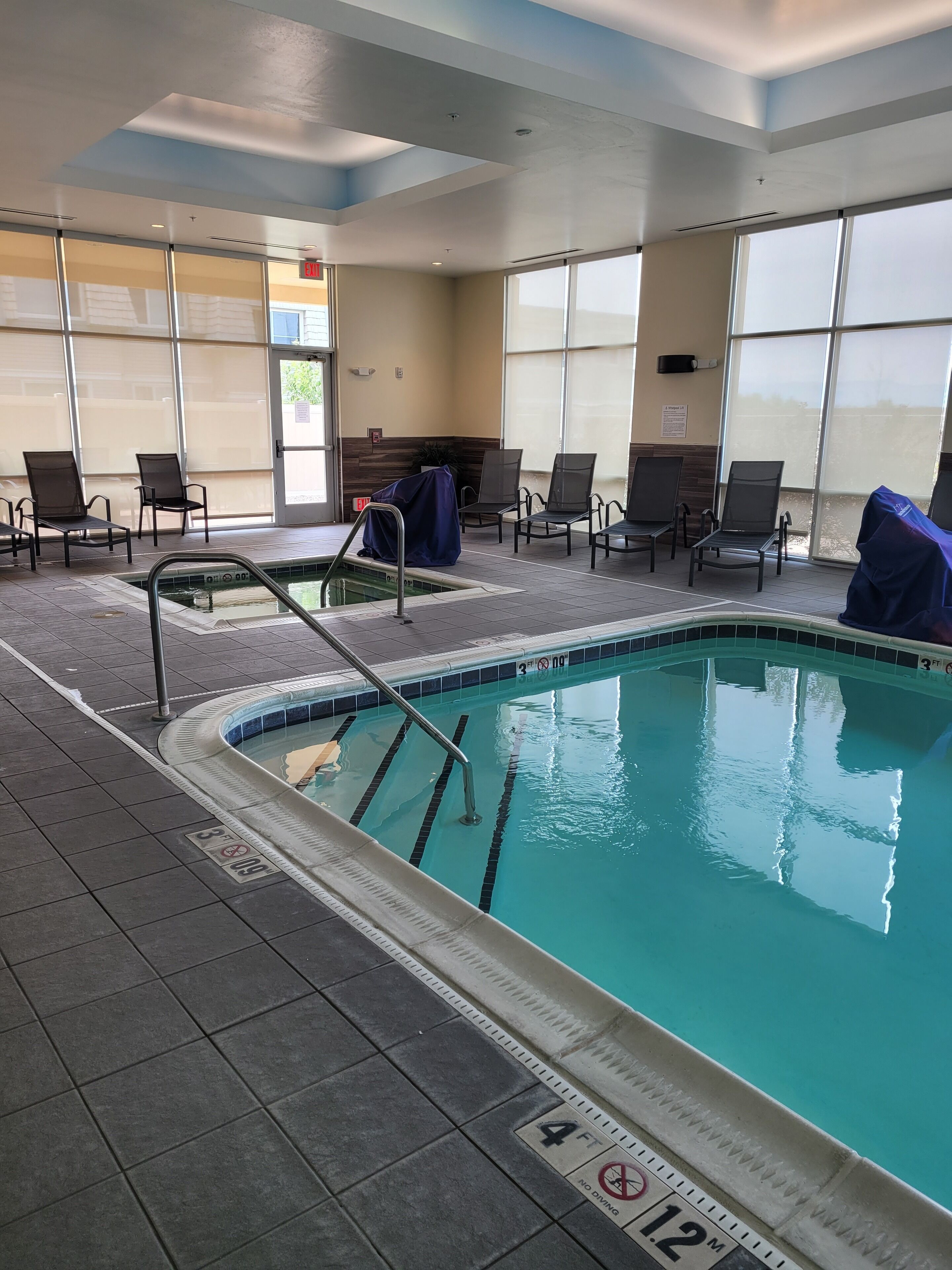 An indoor swimming pool with clear lane markings, a covered seating area with chairs, and a view of the outside through large windows.