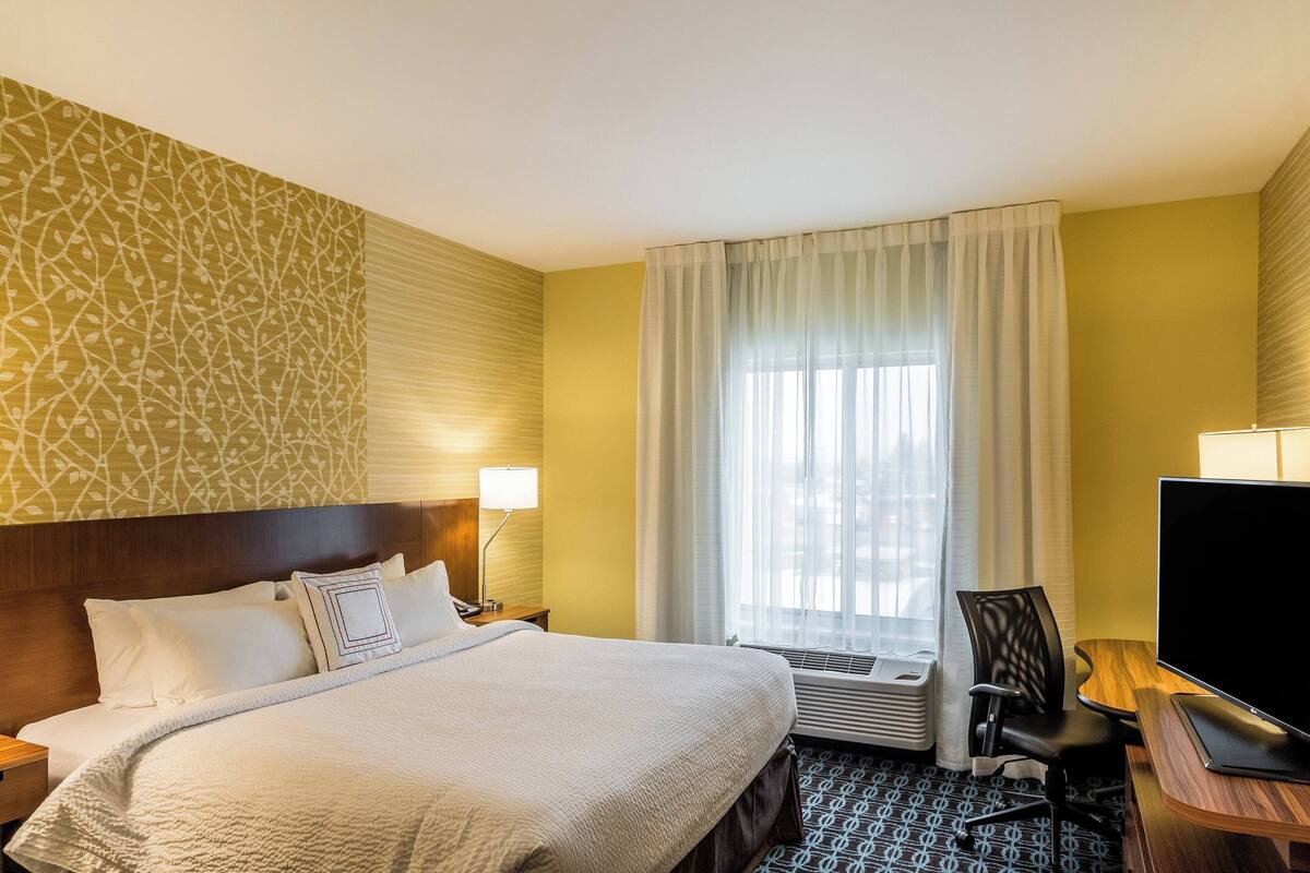 A hotel room with a bed, a desk with a TV, a chair, a window with curtains, and an air conditioning unit.