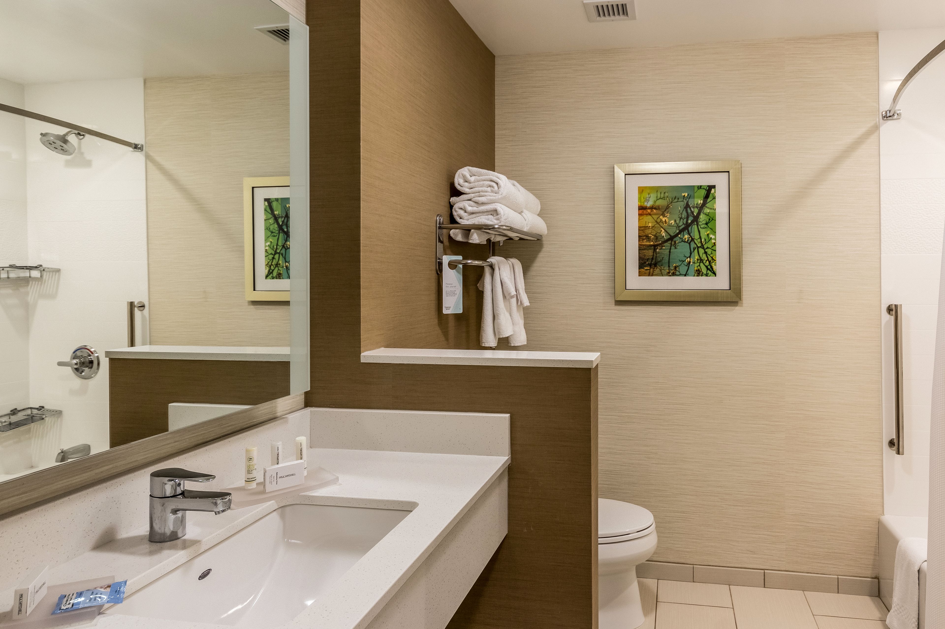 A modern bathroom with a large mirror, a white sink, a toilet, and a shower. There is a towel rack with folded towels and a framed picture on the wall.