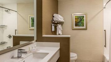 A modern bathroom with a large mirror, a white sink, a toilet, and a shower. There is a towel rack with folded towels and a framed picture on the wall.