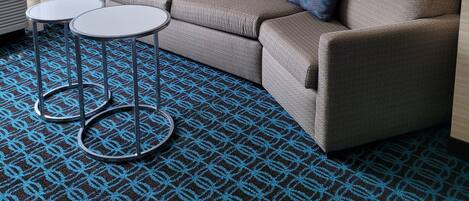 Habitación de hotel con sofá, dos mesitas, un cuadro enmarcado en la pared y una alfombra estampada.