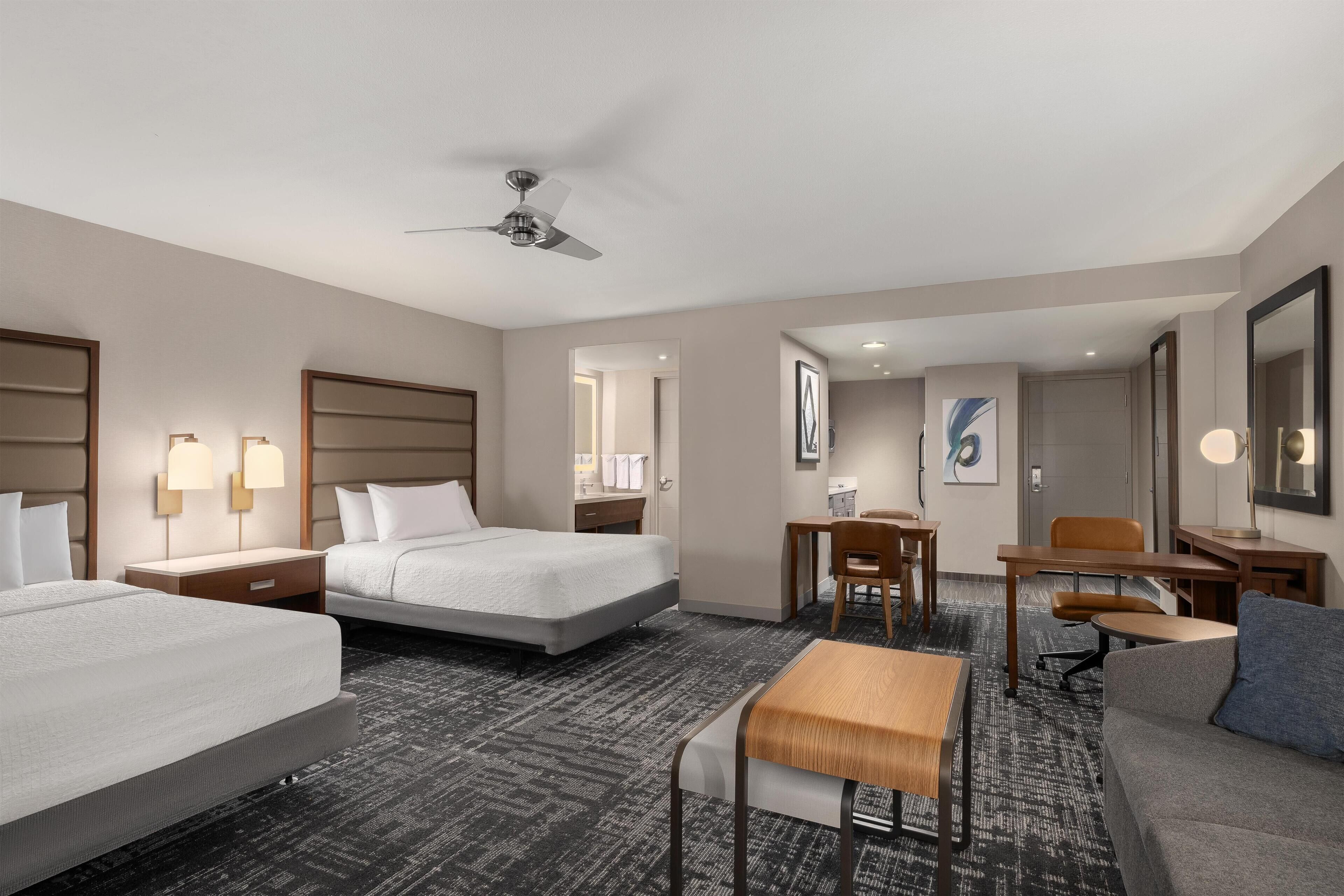 A hotel room with two beds, a desk, a chair, a sofa, a ceiling fan, and a carpeted floor.
