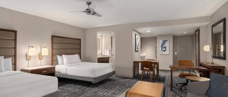 A hotel room with two beds, a desk, a chair, a sofa, a ceiling fan, and a carpeted floor.