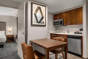Une chambre d’hôtel moderne avec une table à manger en bois, deux chaises, un four à micro-ondes, un lave-vaisselle et une œuvre d’art encadrée au mur.