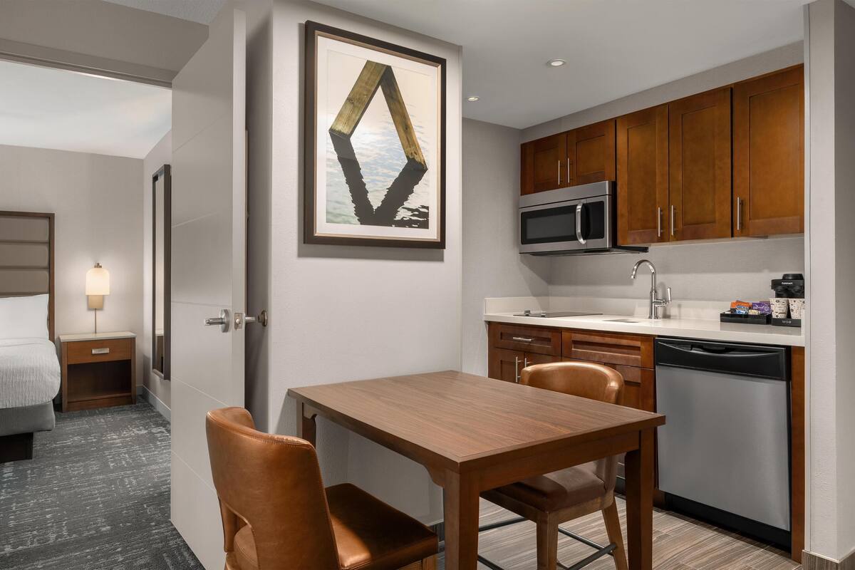 Habitación de hotel moderna con una mesa de comedor de madera, dos sillas, microondas, lavavajillas y una obra de arte enmarcada en la pared.