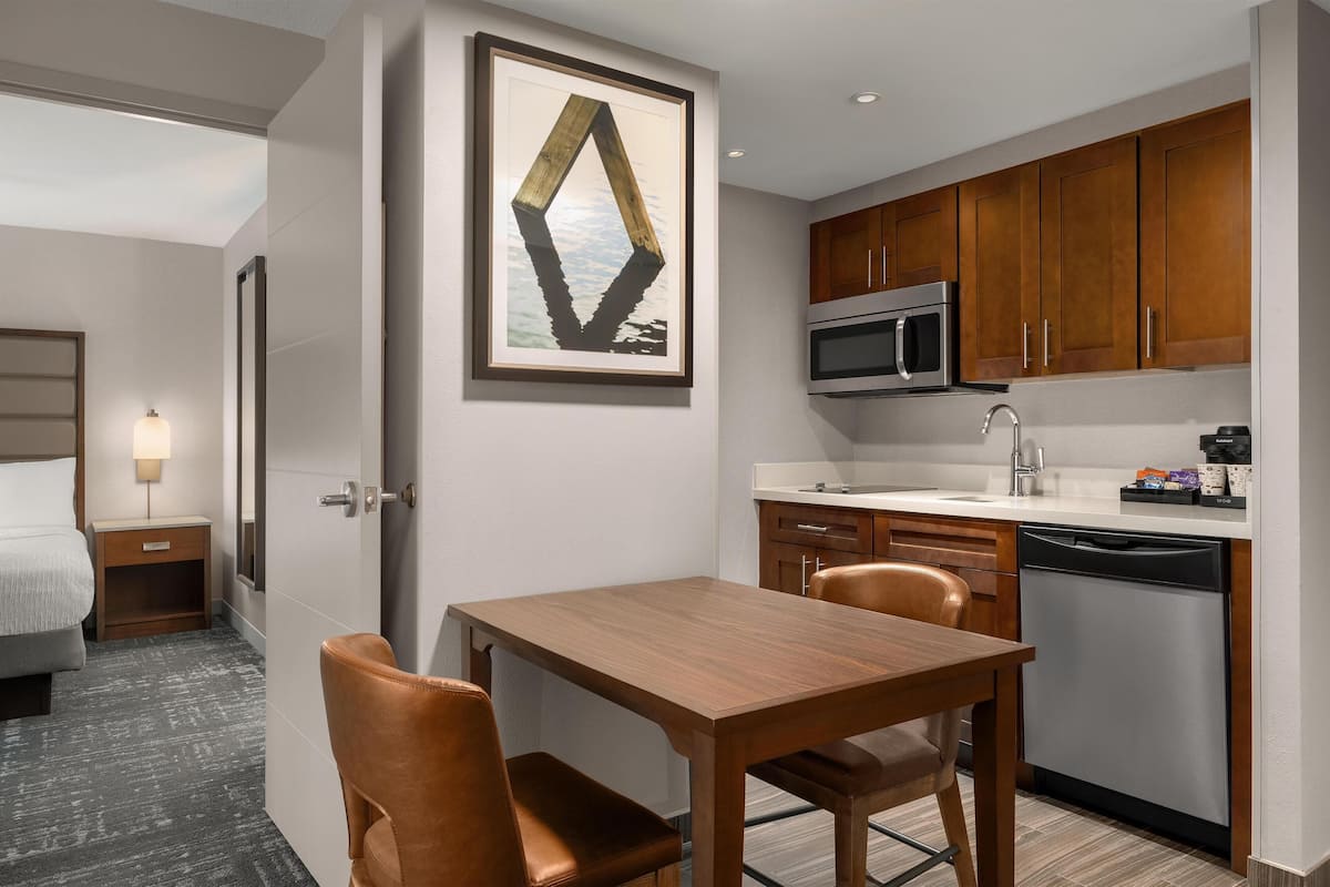 A modern hotel room with a wooden dining table, two chairs, a microwave, a dishwasher, and a framed artwork on the wall.