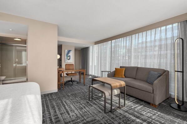 A hotel room with a bed, a sofa, a desk, and a large window with curtains.